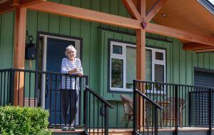 Elder woman stands on porch, built from wood timbers. Modern green siding on house behind her.