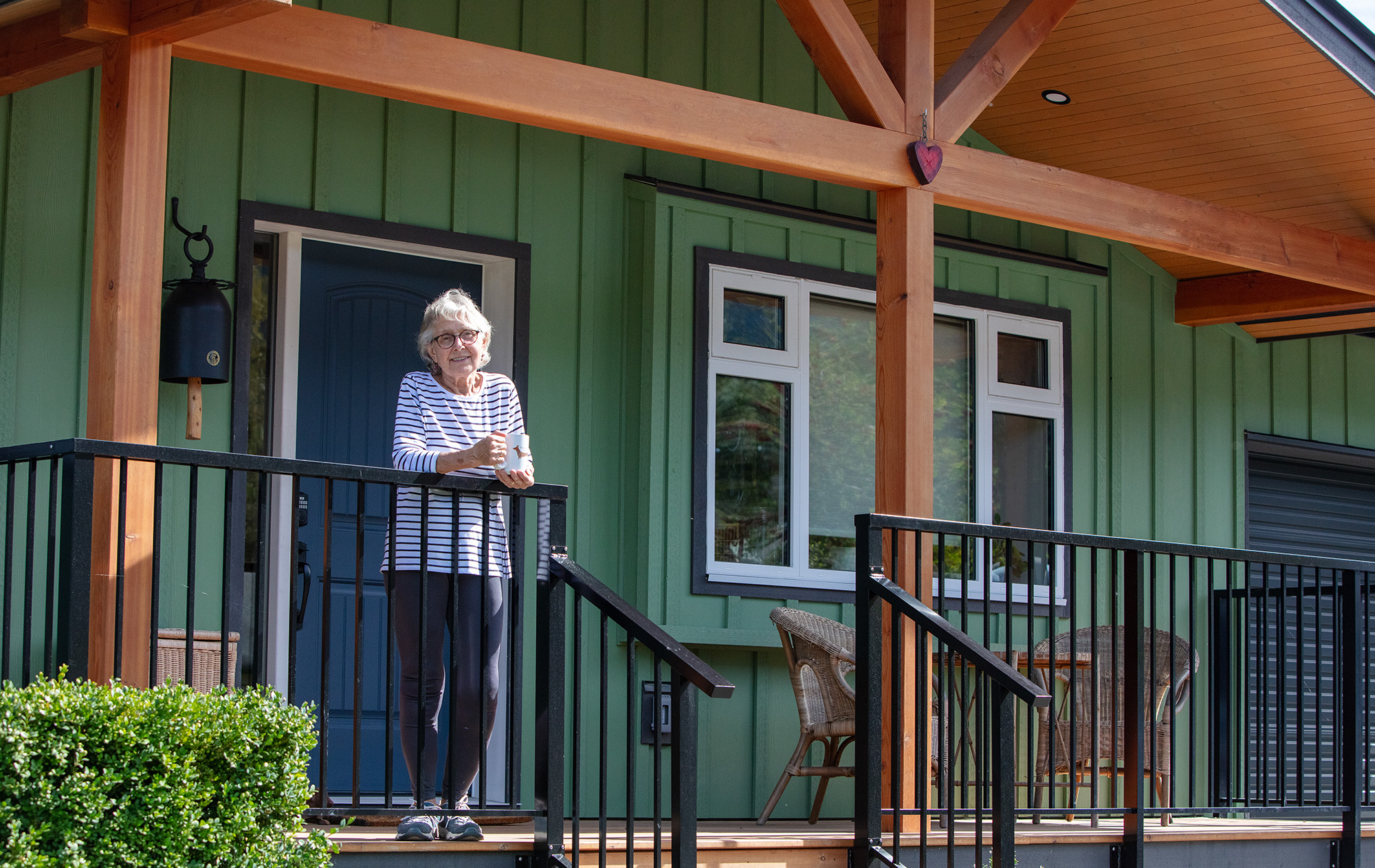 Elder woman stands on porch, built from wood timbers. Modern green siding on house behind her.