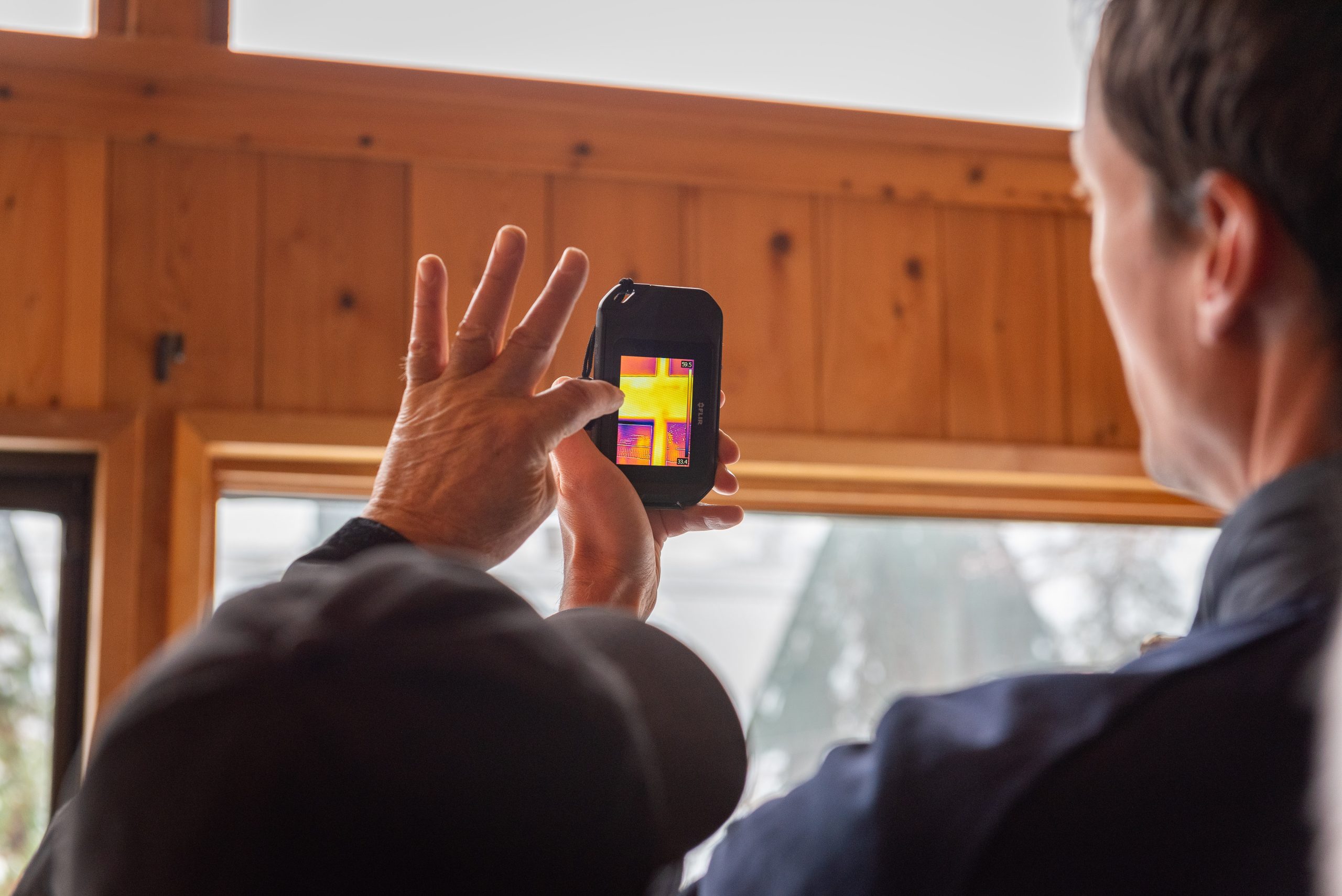 Man holds device showing yellow and orange thermal image of house. Second man is gesturing at device.