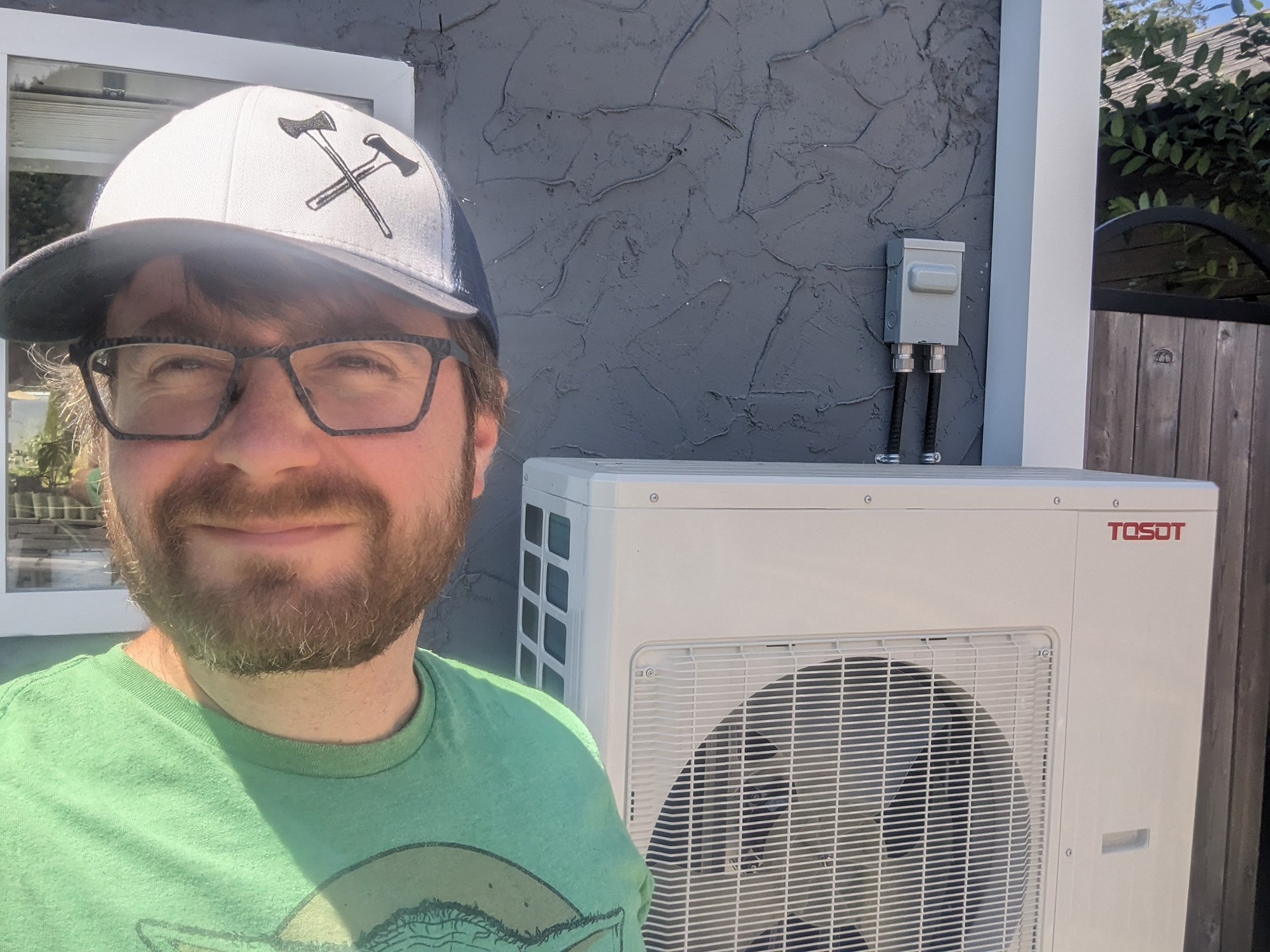 Man in ball cap and glasses smiles at camera. Heat pump on side of home behind him.