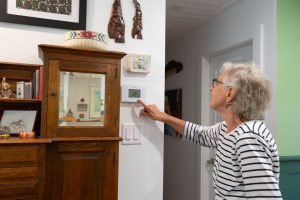 Elder woman presses buttons on electronic thermostat. Surrounded by older, wood furniture.