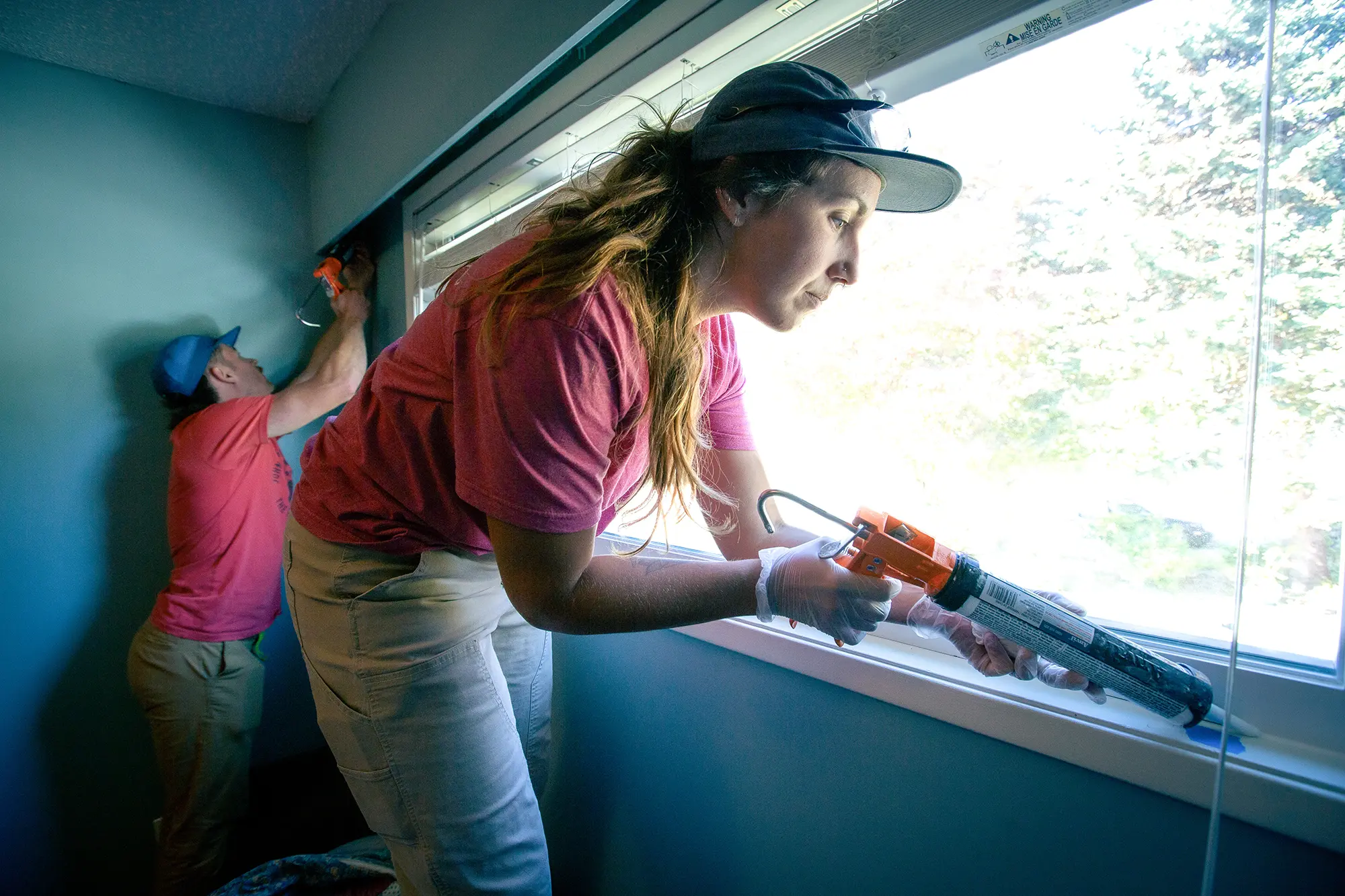 Woman man in red shirts and jeans caulking edges of a large window.