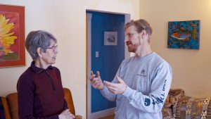 Man in work clothes speaks with elder woman in living room.